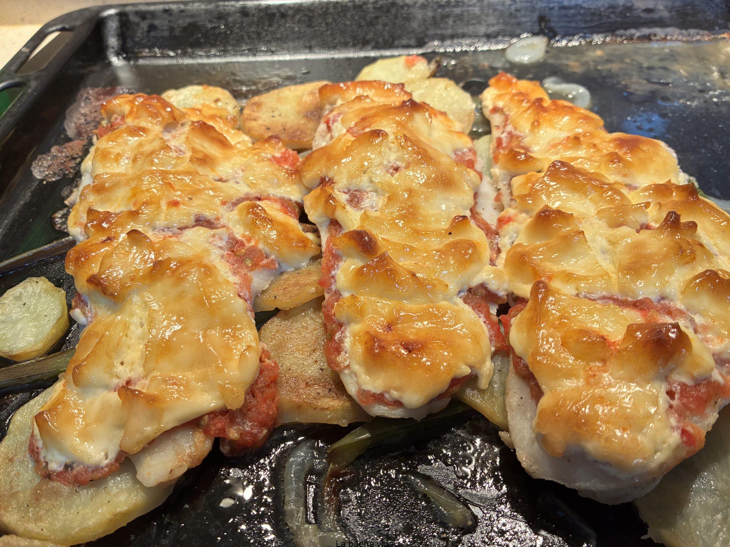 Lomo de merluza horneado con una costra de mahonesa gratinada dorada, servido con patatas panadera y una ensalada fresca con tomates cherry y cebolla roja en un plato rústico.