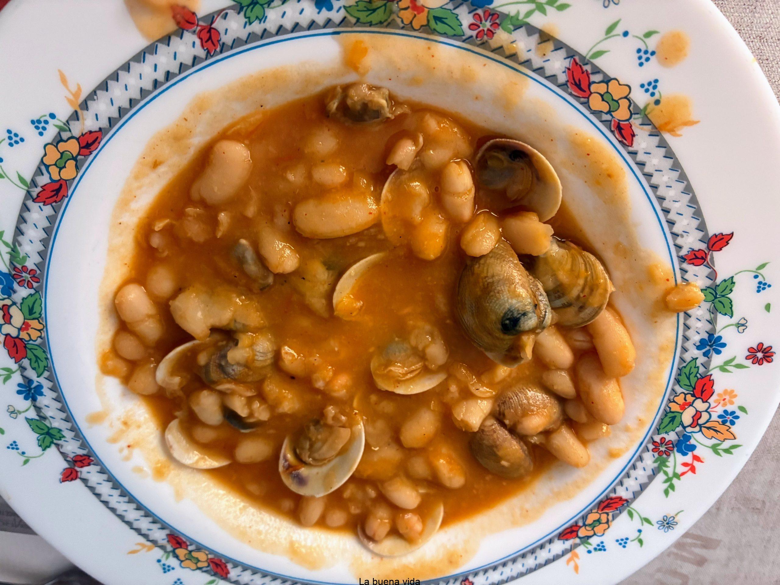 Plato hondo decorado con un guiso caldoso de color anaranjado que contiene alubias blancas enteras y almejas abiertas en su concha.
